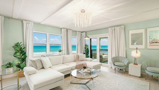 A spacious living room with a large white sofa, a round coffee table, and a view of the ocean through large windows. There are two armchairs and a side table with a lamp.