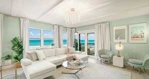 A spacious living room with a large white sofa, a round coffee table, and a view of the ocean through large windows. There are two armchairs and a side table with a lamp. - Auberge Chic - L`Eau: Oceanfront Holiday Escape (St. Augustine Beach)