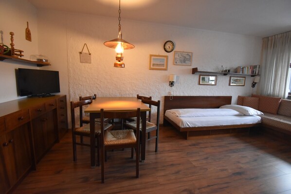 A room with a wooden dining table, chairs, a bed, a television, and a wall-mounted lamp. - NARDIS 205 BILOCALE MhD apt (Madonna di Campiglio)