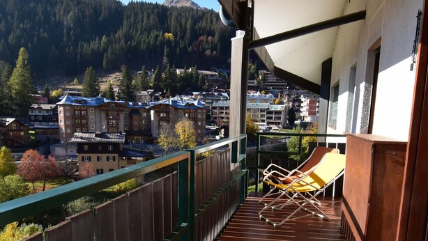 Un balcon doté d’un plancher en bois, d’une chaise jaune et offrant une vue sur une ville et des montagnes en arrière-plan.