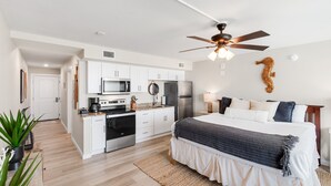 Une chambre moderne avec un grand lit, un ventilateur de plafond et un coin cuisine doté de meubles blancs et d’appareils électroménagers.