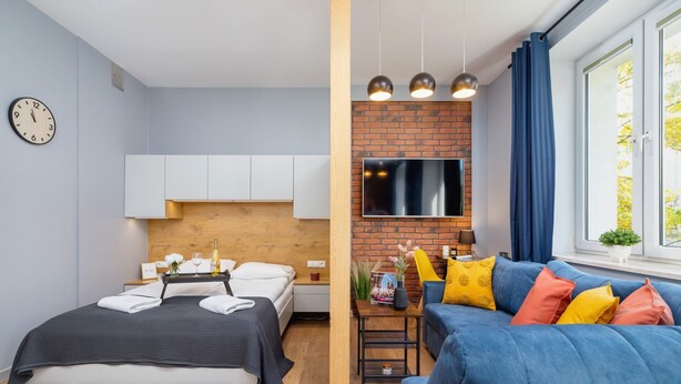 A modern hotel room with a bed, a blue sofa, a TV mounted on a brick wall, and a window with curtains. - Comfortable Studio | Workspace | Close to the Marketplace (Kraków)