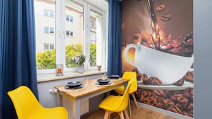 A dining area with a table set for two, yellow chairs, a window with blue curtains, and a mural of coffee beans on the wall. - Comfortable Studio | Workspace | Close to the Marketplace (Kraków)