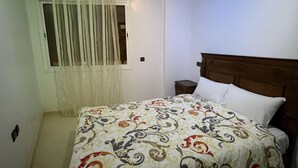 Une chambre à coucher comprenant un lit, une tête de lit en bois, une table de chevet, une fenêtre avec des rideaux et une porte.