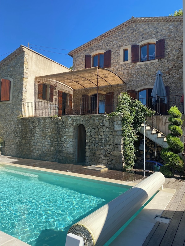 A stone building with a pool, a covered patio, and a staircase leading to a balcony.