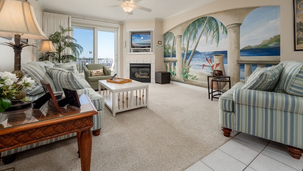 A spacious living room with striped sofas, a fireplace, a ceiling fan, and a large mural of a beach scene. - Beautiful 3 Bedroom, 3 Bath Oceanblock Condo Mid-Town with Indoor Pool (Ocean City)
