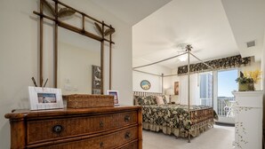 A bedroom with a four-drawer wooden dresser, a canopy bed with floral bedding, a large mirror, and a view of the outdoors through a balcony. - Beautiful 3 Bedroom, 3 Bath Oceanblock Condo Mid-Town with Indoor Pool (Ocean City)
