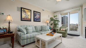 A living room with a striped sofa, a white coffee table, a green armchair, and a large window with a city view. - Beautiful 3 Bedroom, 3 Bath Oceanblock Condo Mid-Town with Indoor Pool (Ocean City)
