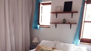 A bedroom with a bed, white curtains, a window with blue drapes, and wall-mounted shelves with decorative items. - Casa Balcón- Increíbles Vistas! (Saleres)