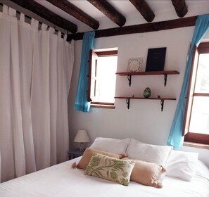 Classic bedroom with a bed, white linens, and decorative pillows, set against a wooden ceiling with exposed beams and a window with curtains.