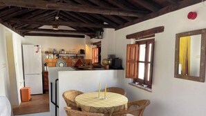 A rustic kitchen with wooden beams, a round table with wicker chairs, and a window with a yellow curtain. - Casa Balcón- Increíbles Vistas! (Saleres)