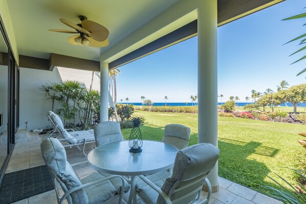 A covered patio with a round table and chairs, a ceiling fan, and a view of the ocean. - Enjoy Ocean Views from Mauna Lani Point A105 (Kamuela)