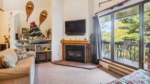 Uma sala de estar com lareira, sofá, televisão e uma grande janela com vista para árvores.