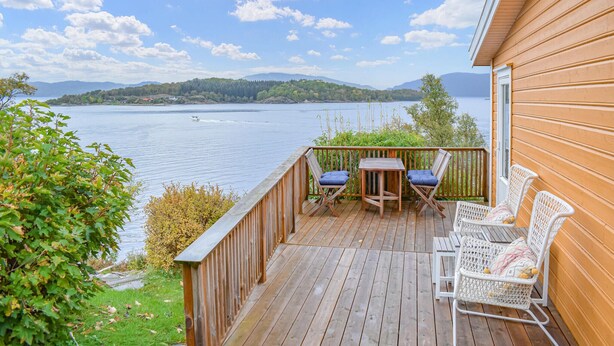 Une terrasse en bois avec une table et des chaises, donnant sur un lac et offrant une vue sur des montagnes au loin.