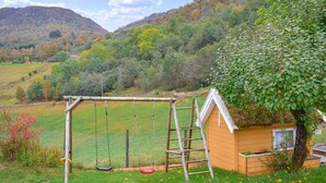 Une cabane en bois, avec un portique et une échelle, se trouve dans une cour verdoyante, offrant une vue sur une colline boisée.