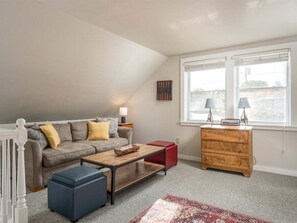 A loft-style room with a high ceiling, a sofa set, a wooden coffee table, a red ottoman, a wooden dresser, and a window with blinds. - Bethel Village Carriage House Apartment (Bethel)