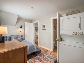 A bedroom with a bed, a wooden dresser, a washing machine, and a door leading to another room. - Bethel Village Carriage House Apartment (Bethel)