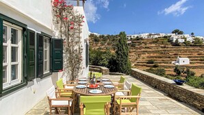 A patio with a round table set for dining, surrounded by chairs, with a view of terraced fields and white buildings in the background. - Gorgeous home in Sifnos (Sifnos)