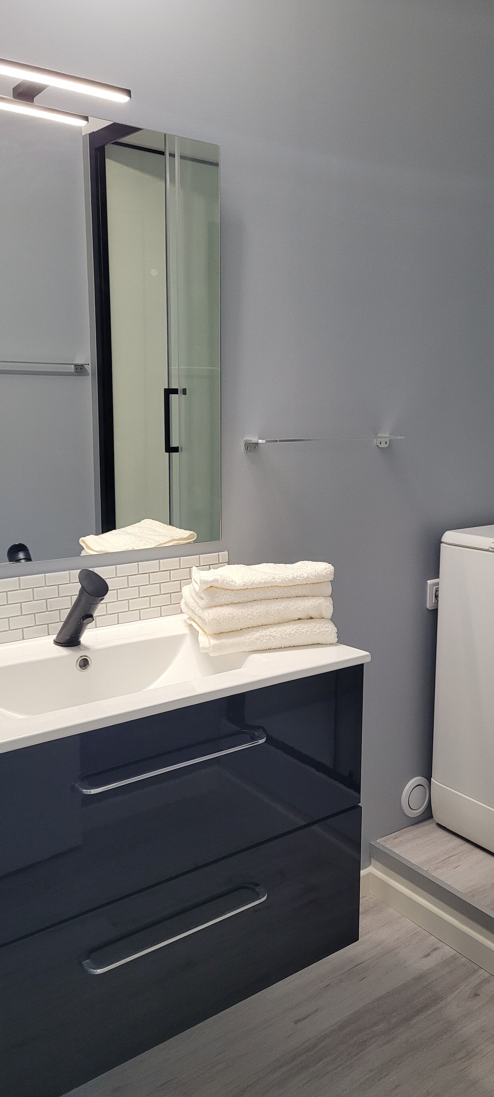Ein modernes Badezimmer mit einem weißen Waschbecken, einem schwarzen Schrank und einem Spiegel darüber. Gefaltete Handtücher liegen auf dem Waschbecken, und eine Toilette ist rechts im Bild zu sehen.
