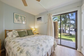 Ein Schlafzimmer mit einem Bett, einem Nachttisch, einer Wand-Klimaanlage und Blick auf Palmen durch eine Schiebetür.
