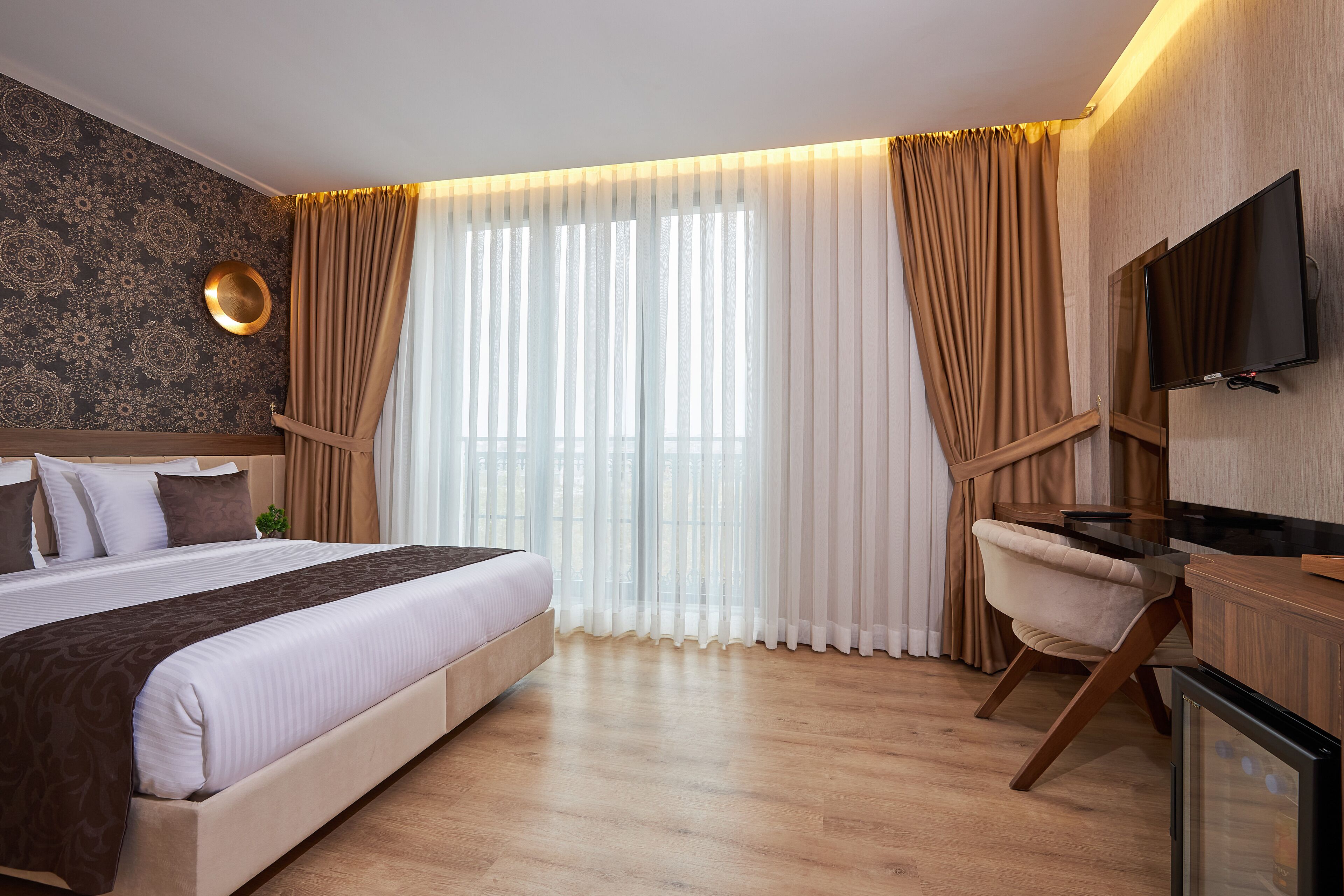 A modern hotel room with a large bed, a desk, a television, and a view of the outdoors through sheer curtains.
