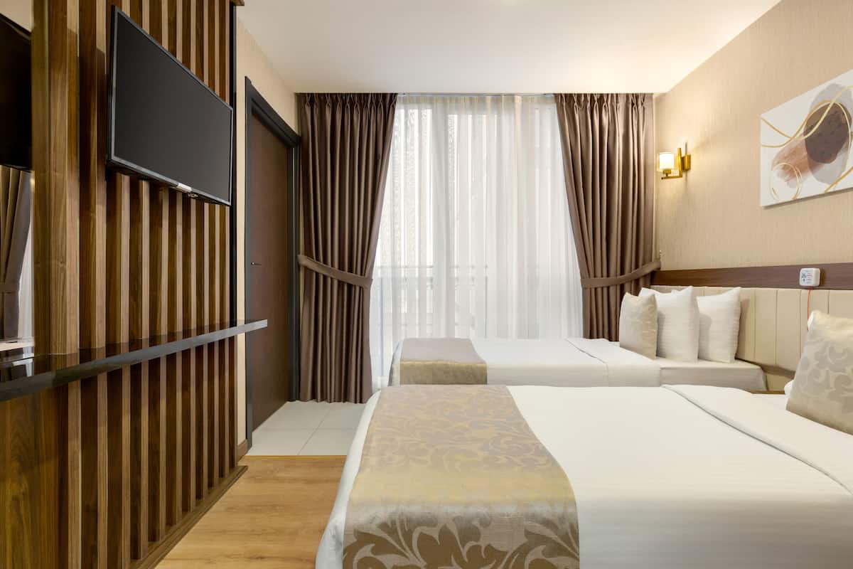 A hotel room with two beds, a flat-screen TV mounted on a wooden panel, a large window with sheer curtains, and a wall-mounted light fixture.