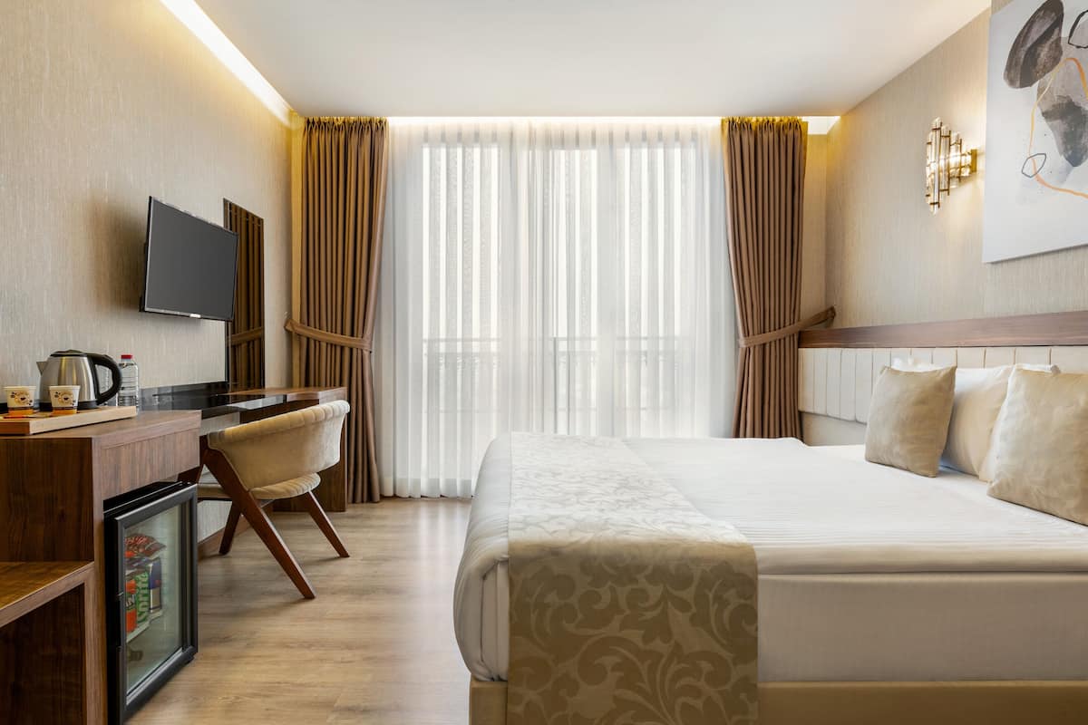 A hotel room with a bed, a desk with a television, a mini-fridge, and a window with curtains.