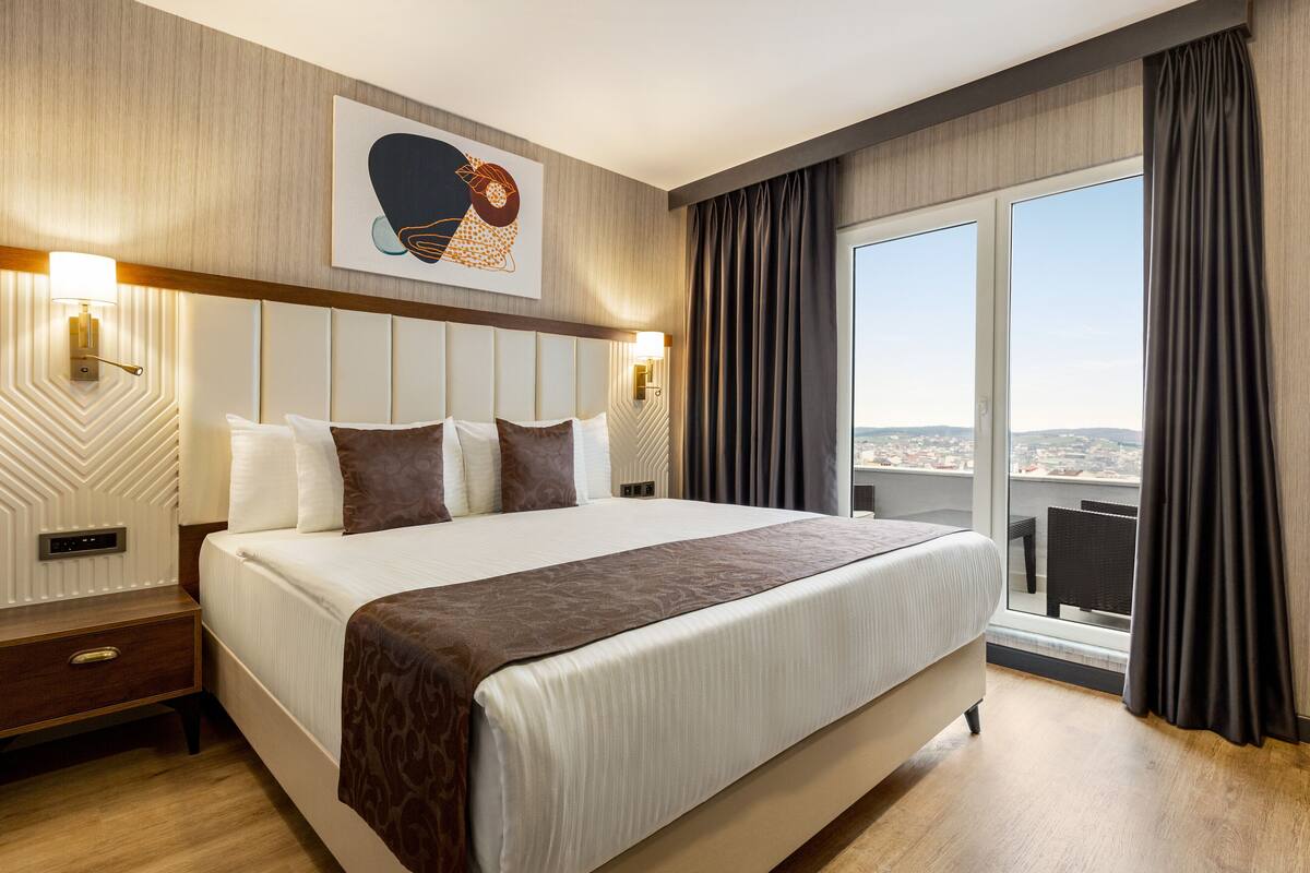 A modern hotel room with a large bed, a city view, and a decorative wall piece.