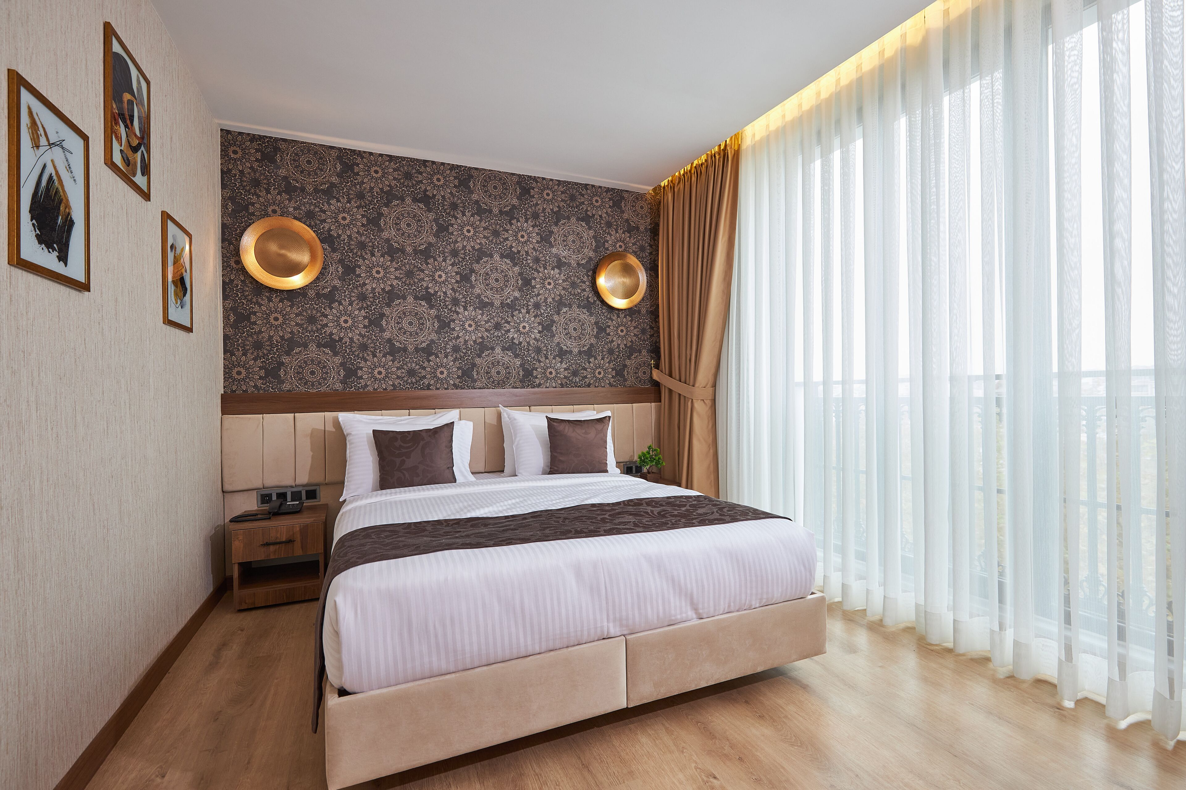A modern hotel room with a large bed, a nightstand, a wall-mounted lamp, and a view of the outdoors through sheer curtains.