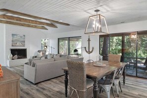 A modern living room with a fireplace, a dining area with a wooden table and chairs, and large windows offering a view of the outdoors.