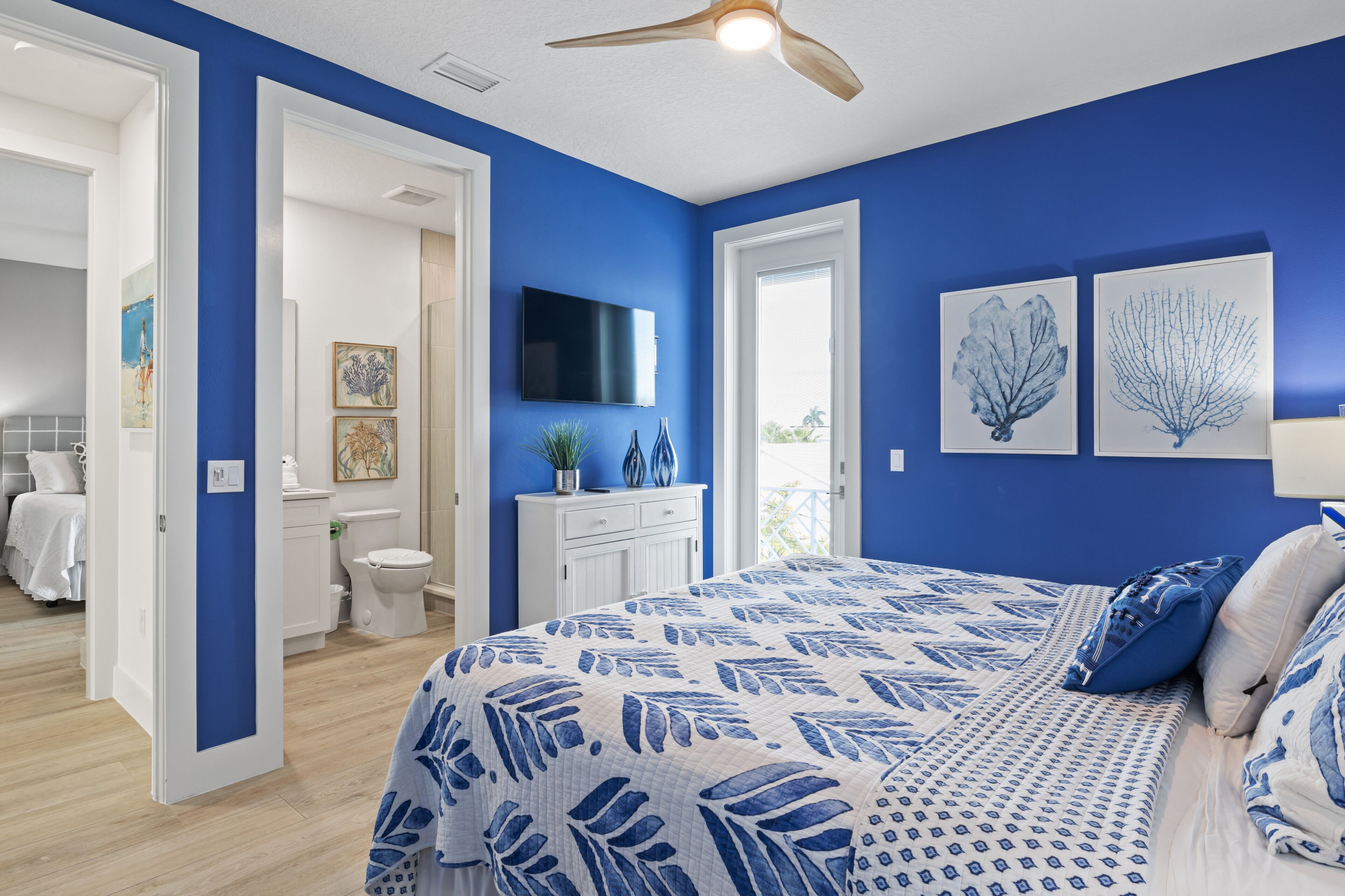 A bedroom with a blue accent wall, a bed with a leaf-patterned duvet, a ceiling fan, and artwork on the wall.