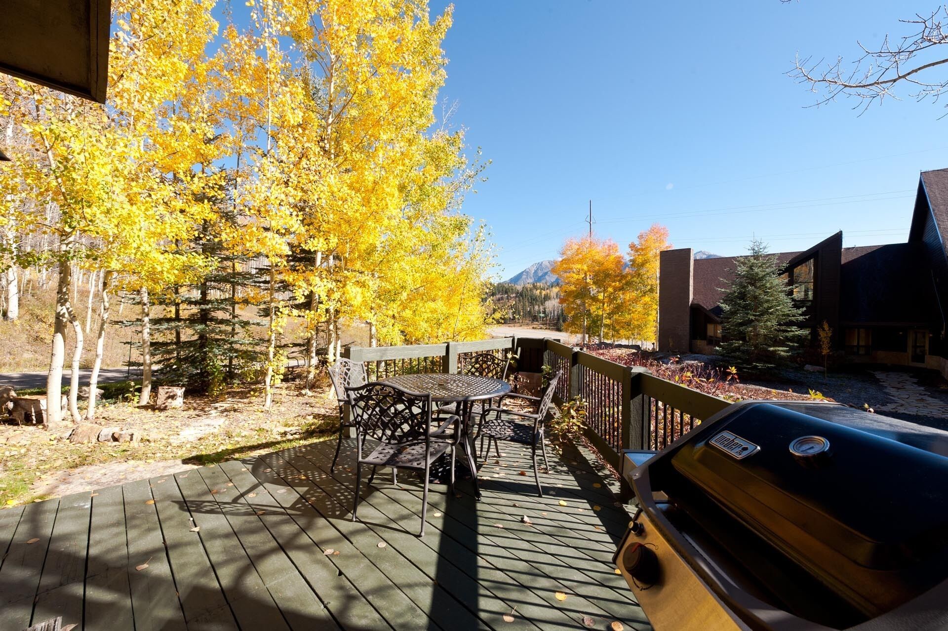 A deck with a grill, a table, and chairs, surrounded by trees with yellow leaves.