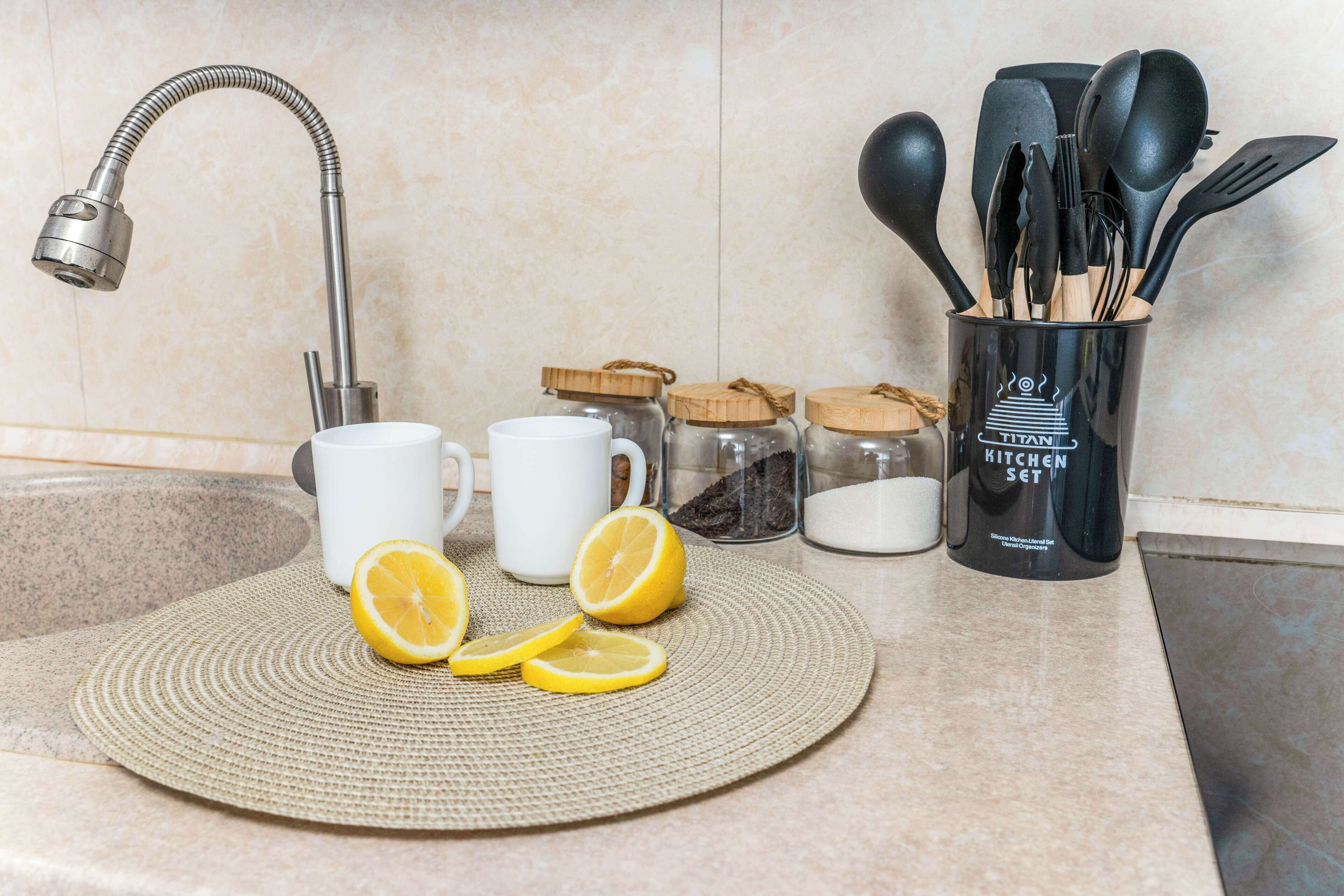 Una encimera de cocina con un grifo de acero inoxidable, tazas blancas, rodajas de limón, un posavasos tejido y un recipiente negro para utensilios con la etiqueta 'TITAN KITCHEN SET'.