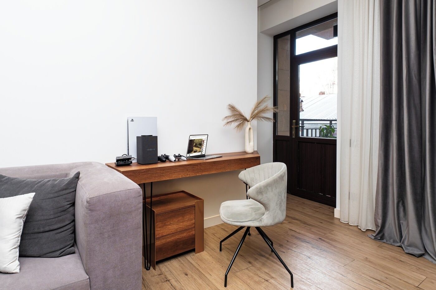 Una sala de estar moderna con un sofá gris, un escritorio de madera con una computadora portátil, una consola de videojuegos y una silla gamer.