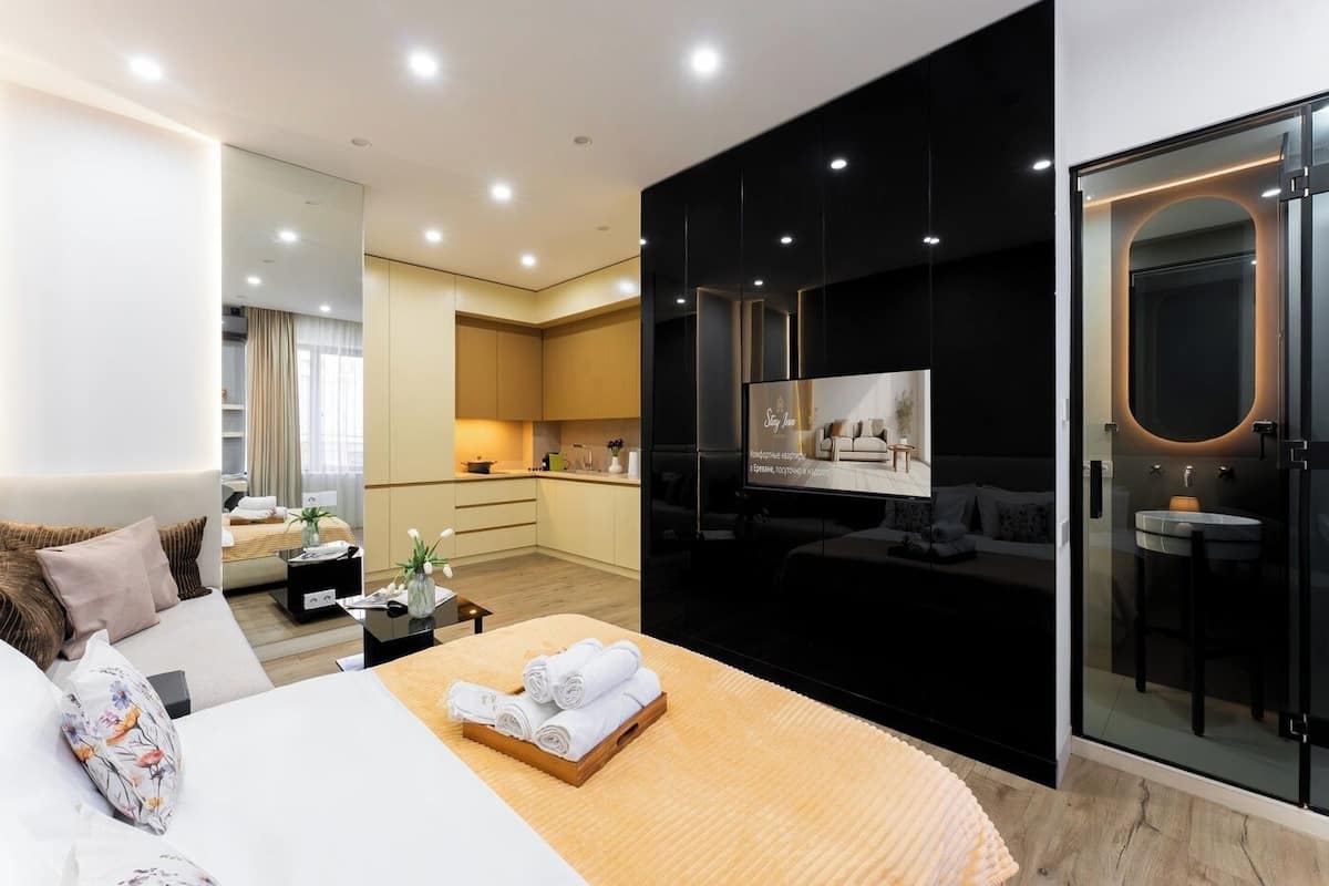 Een moderne hotelkamer met een bed, nachtkastje met handdoeken, een klein tafeltje met een vaas, een grote spiegel, een televisie en een badkamer met een wastafel en spiegel.