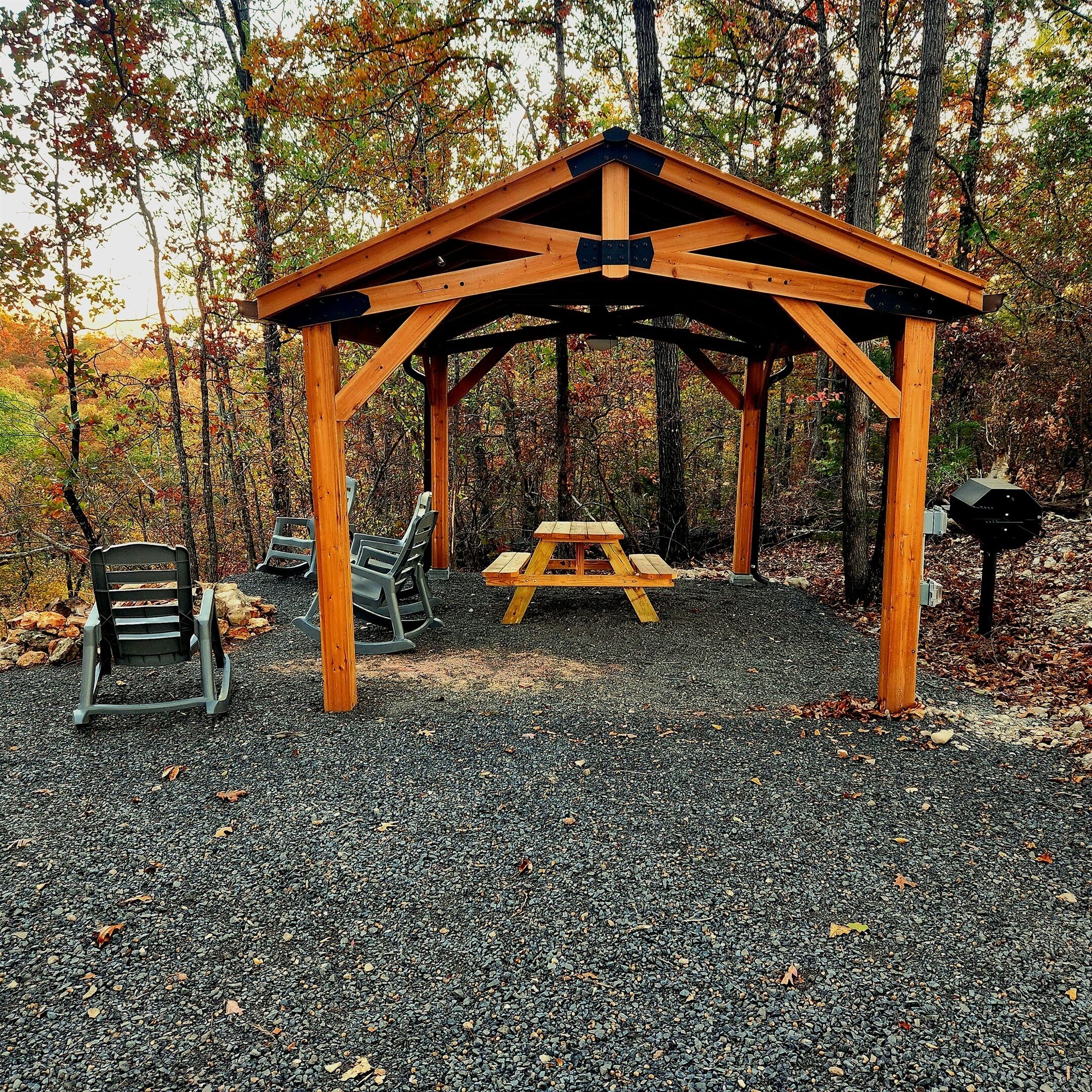 A wooden pavilion with a picnic table and two rocking chairs in a forested area.
