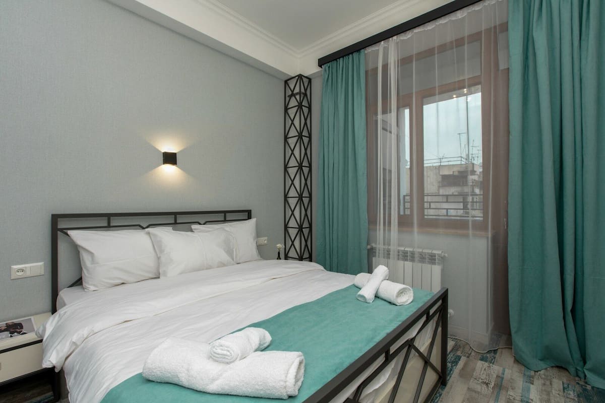 A hotel room with a bed, a nightstand, a wall-mounted light, a window with curtains, and a view of buildings outside.