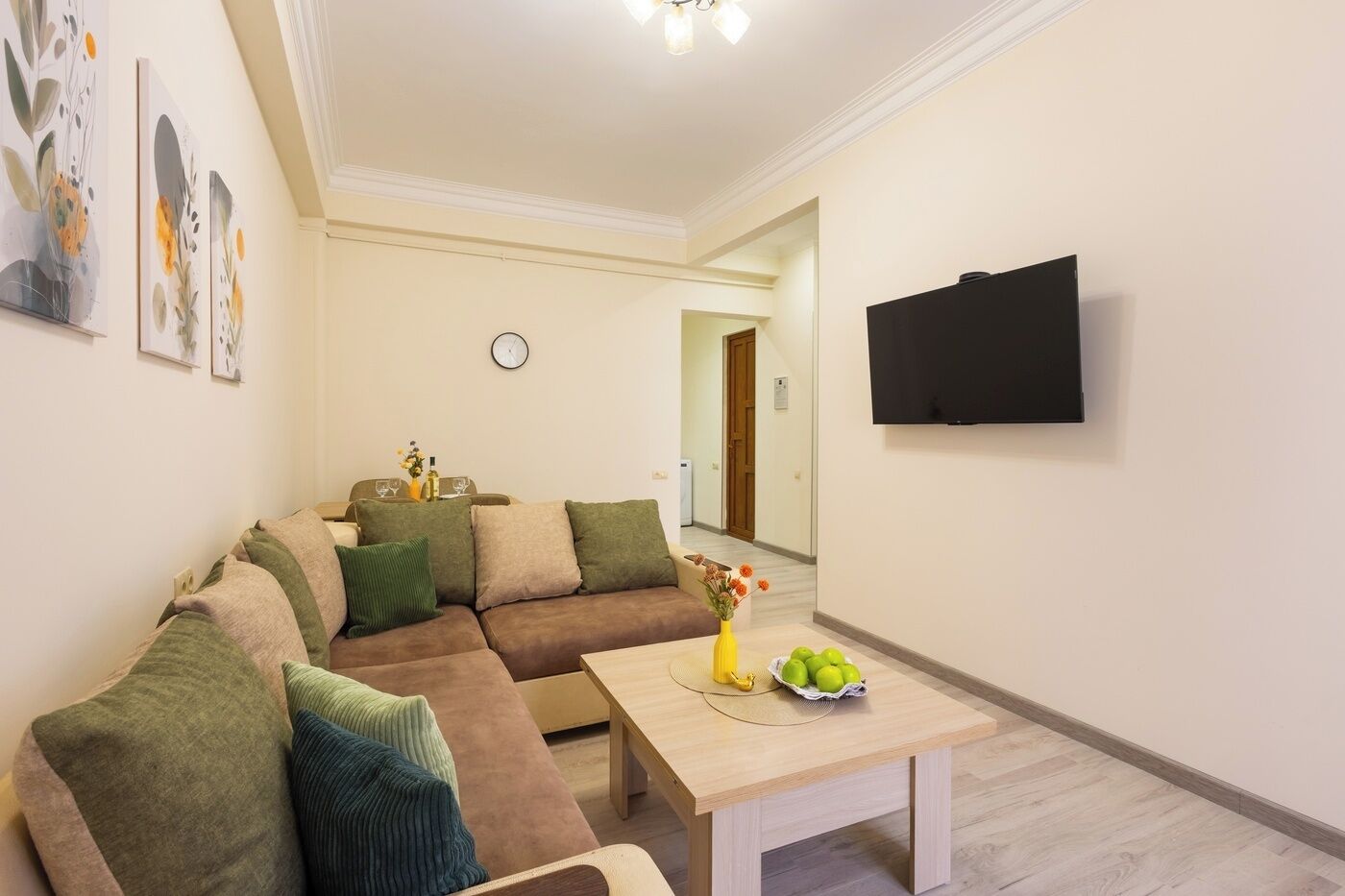 A living room with a sectional sofa, a coffee table with fruit, a flat-screen TV, and a clock on the wall.