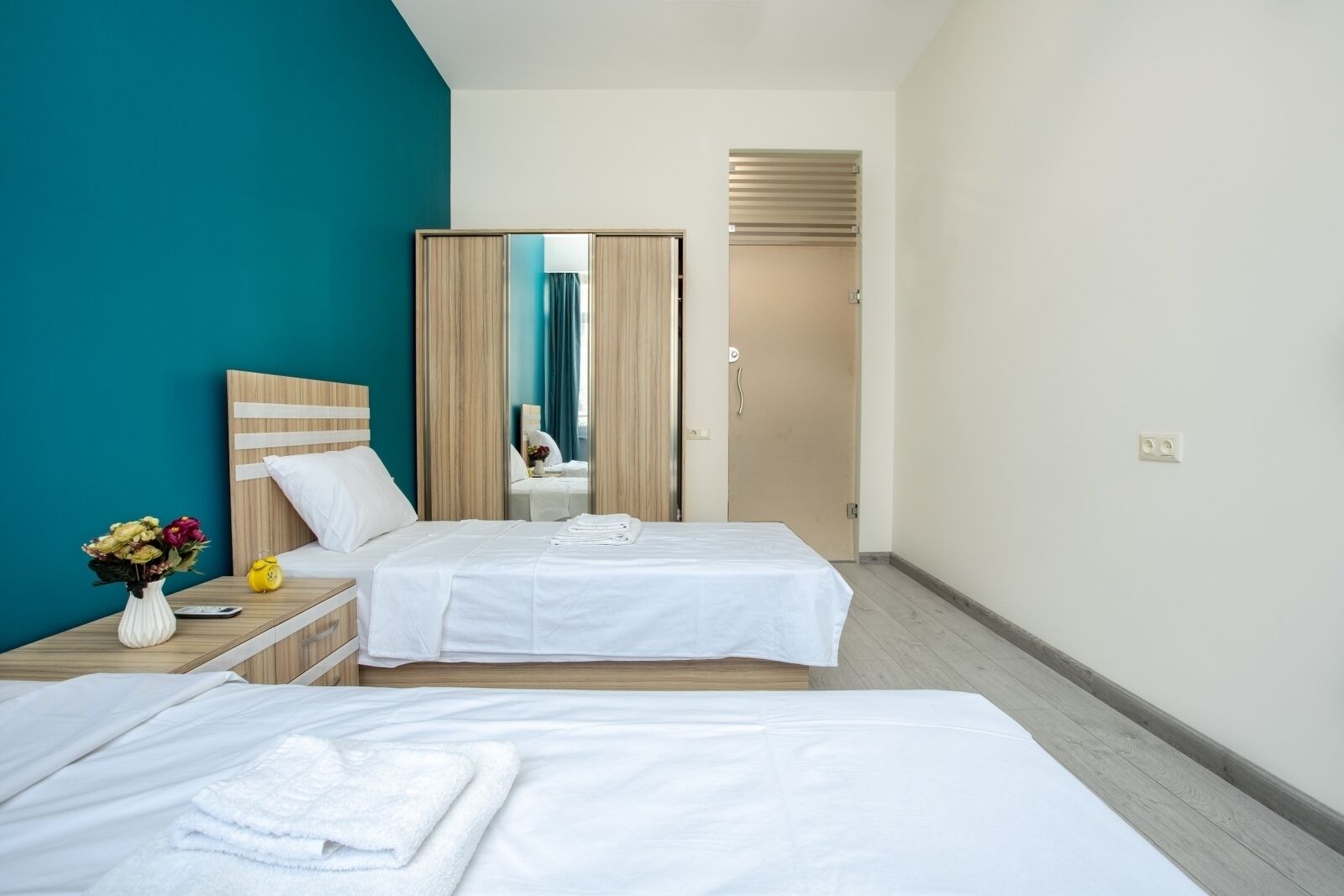 Habitación de hotel con dos camas, una mesita de noche, un jarrón con flores y vista al baño a través de un espejo.