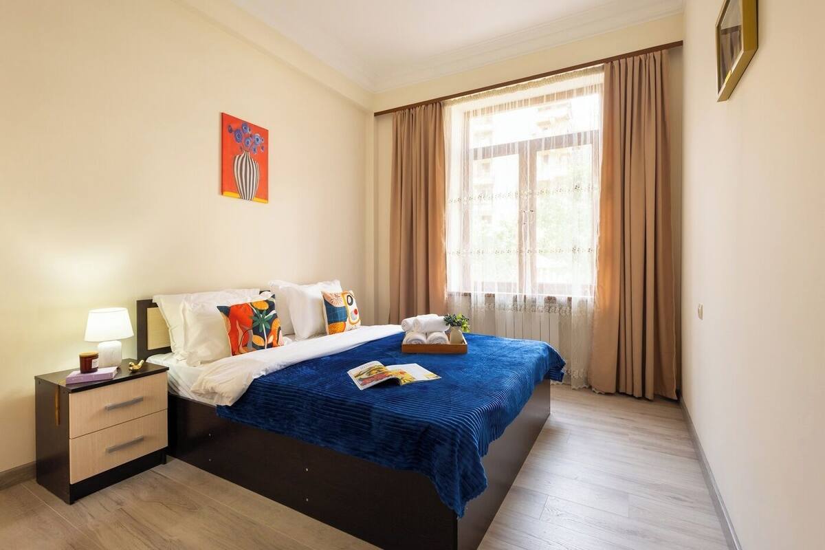 A hotel room with a bed, a nightstand, a lamp, a window with curtains, and a painting on the wall.