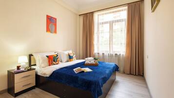 Habitación de hotel con cama, mesita de noche, lámpara, ventana con cortinas y un cuadro en la pared.