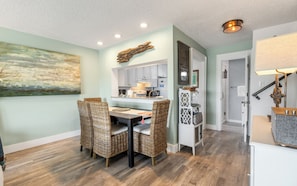 A dining area with wicker chairs, a kitchen with light-colored cabinets, and a hallway with a door leading to a bathroom. - Inlet Beach + Gulf View, Pet Friendly & Bikes (Inlet Beach)