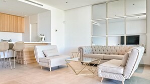 Ett modernt vardagsrum med en soffa, fåtöljer och ett soffbord. Här finns också en köksdel med ett handfat och en symaskin.