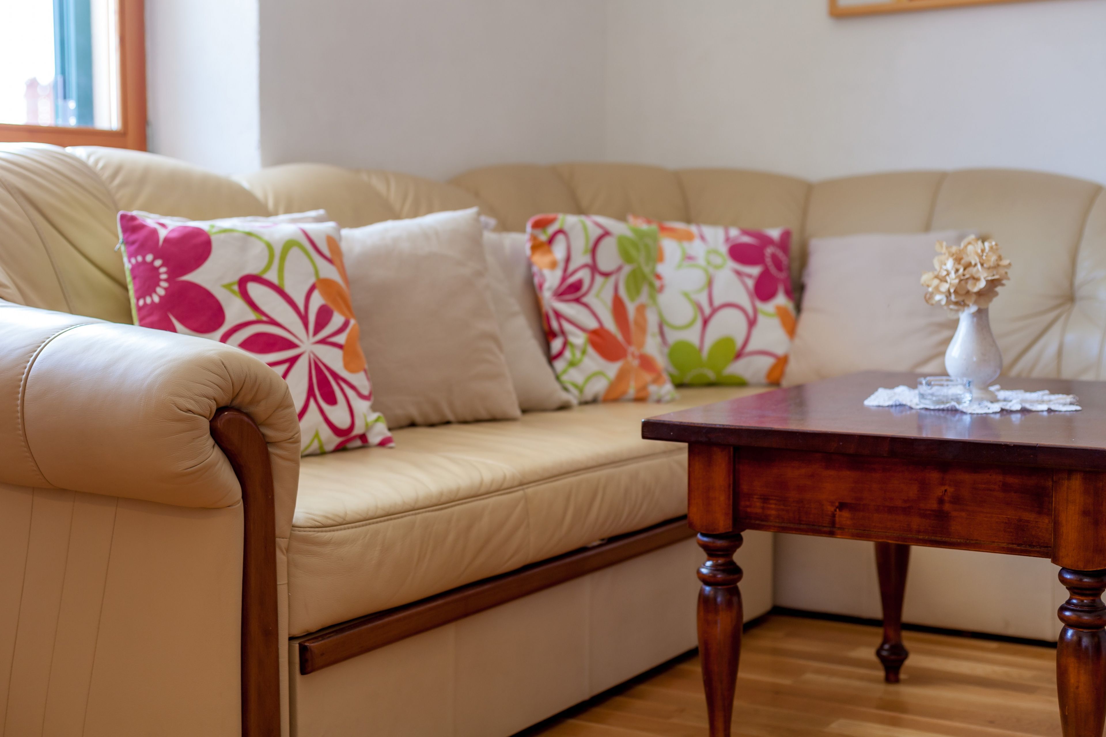A beige leather sofa with colorful floral cushions, a wooden table with a vase, and a wooden floor.