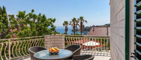 Un balcón con mobiliario de mimbre, una mesa con frutero y vistas al mar y palmeras.