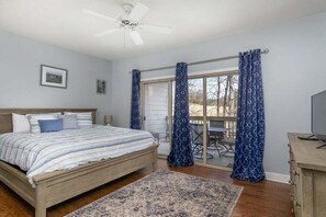 A bedroom with a bed, a ceiling fan, a large window with curtains, and a view of the outdoors.