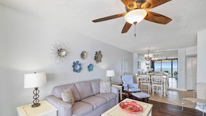 A living room with a beige sofa, white side tables, a ceiling fan, and decorative wall art.