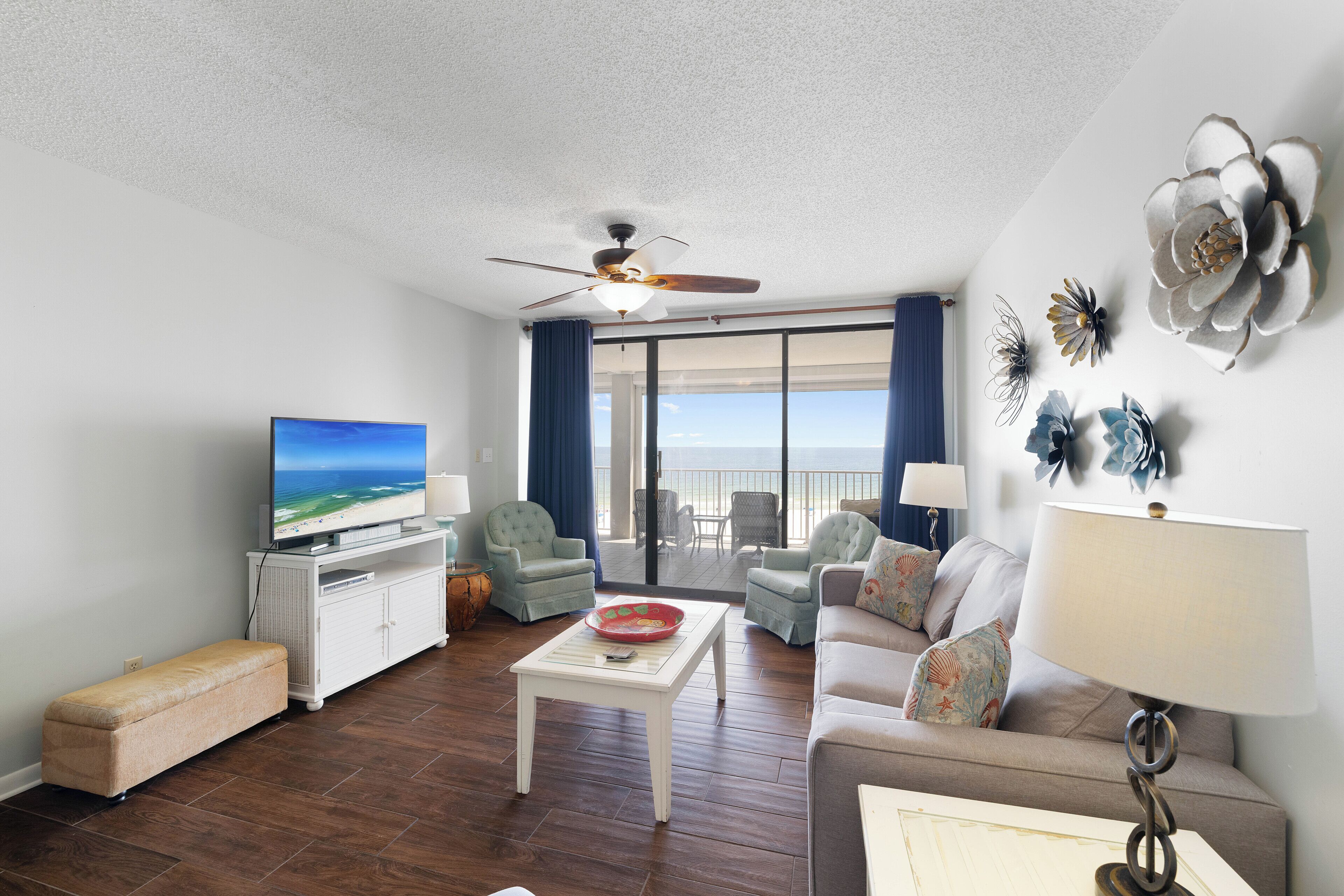 Ein Wohnzimmer mit Fernseher, Deckenventilator, Sofa, Couchtisch und Meerblick durch große Fenster.