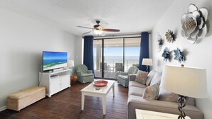 A living room with a television, a ceiling fan, a sofa, a coffee table, and a view of the ocean through large windows.
