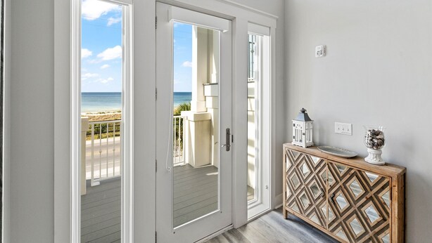 A room with a view of the beach, a wooden console table with decorative items, and a white door leading to an outdoor balcony.